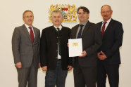 Vier Personen vor dem Bayerischen Staatswappen mit Ernennungsurkunde