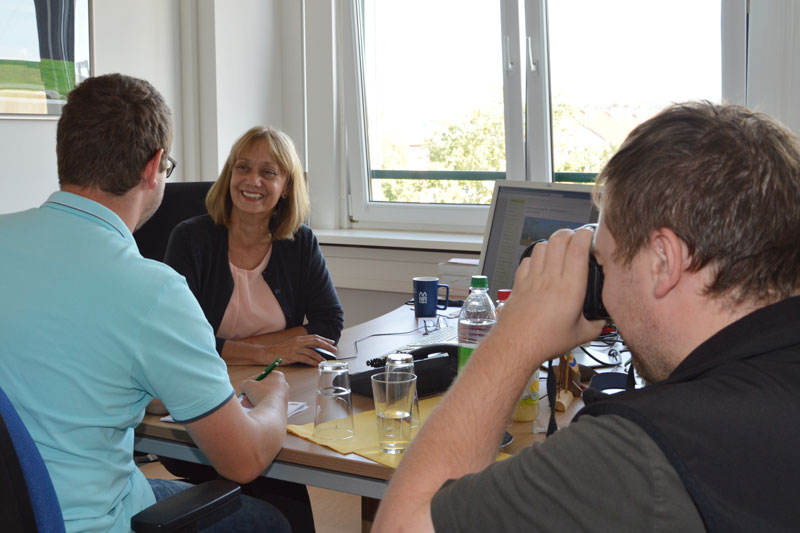 Zwei Personen sitzend am Tisch, eine Person im Vordergrund fotografiert knieend 